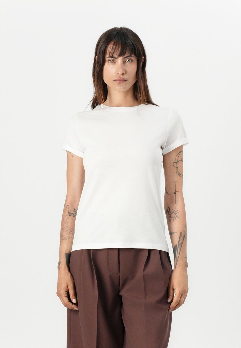 White cotton t-shirt with short rolled sleeves, rounded neck, and a straight hem. Worn with brown loose trousers. Model stands against a white background.