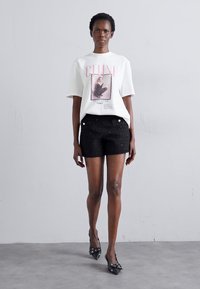 T-shirt graphique blanc avec le texte "Celine" en rose, associé à un short noir texturé avec des accents de boutons argentés et des talons aiguilles noirs.
