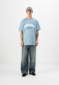 Ung mand står, iført en lys blå Jack & Jones T-shirt, brede falmede jeans, sorte sneakers og en grå kasket, mod en helt hvid baggrund.