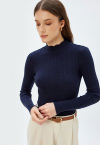 Navy blau gerippter Strickpullover mit einem gewellten hohen Kragen und langen Ärmeln, kombiniert mit einem hellbraunen Gürtel und cremefarbenen Hosen.