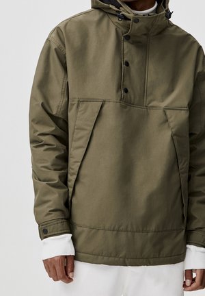 Personne portant une veste à capuche vert olive avec des boutons noirs et des manches blanches superposées, vue des épaules jusqu'aux hanches.