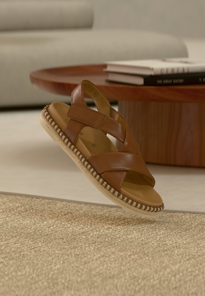 Sandale en cuir marron avec des sangles croisées, une semelle intérieure rembourrée et une semelle tressée, posée sur un fond de tapis tissé clair.