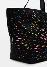 Black tote bag with cut-out design revealing colorful interior. Textured material; features a logo tag on the side. Handles for carrying.