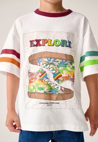 Fehér póló, amelyen színes, terepmintás tornacipő illusztráció látható, borvörös nyakkal és zöld valamint narancssárga csíkos díszítéssel.