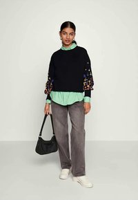 Γυναίκα με μαύρη μπλούζα με πολύχρωμες λουλουδάτες κεντήσεις στα μανίκια, μέντα πράσινο πουκάμισο, γκρι παντελόνι, λευκά παπούτσια, και κρατώντας μια μαύρη τσάντα.