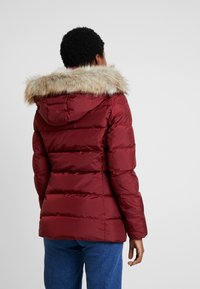 Person som bär en vinröd vadderad jacka med huva kantad med päls, stående med ryggen mot kameran mot en neutral bakgrund.