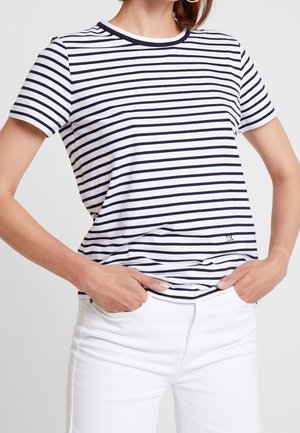 Femme les mains dans les poches, portant une chemise à manches courtes rayée blanche et marine et un pantalon blanc, sur un fond uni.