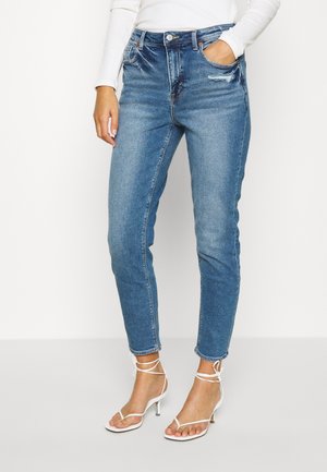 Frau in blauen, schmal geschnittenen Jeans mit leichter Used-Optik, weißem Langarmshirt und weißen, mit Riemchen versehenen Sandaletten mit Absatz, stehend vor weißem Hintergrund.