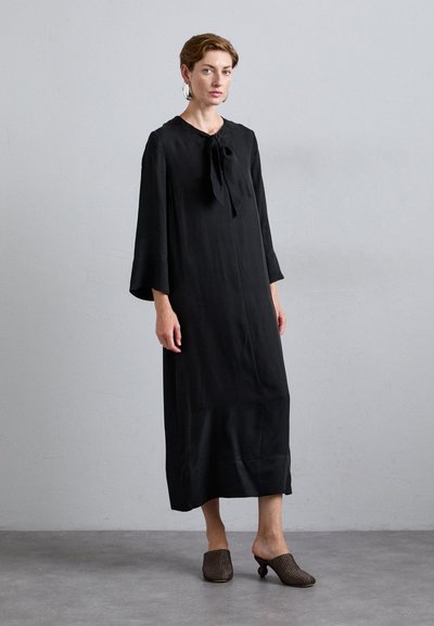 Black, long-sleeved dress with a tie neckline and straight silhouette. The model wears textured brown slip-on shoes. Simple design and smooth fabric.
