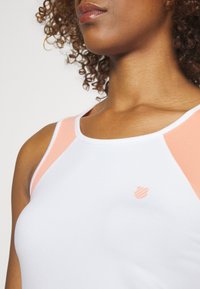 Femme portant un haut blanc sans manches avec des panneaux rose clair et un petit logo en forme de bouclier rose sur la poitrine, cheveux bouclés visibles.