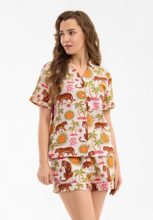 Femme portant un ensemble chemise à manches courtes boutonnée et short avec un imprimé coloré de tigre, soleil et palmier sur fond clair.