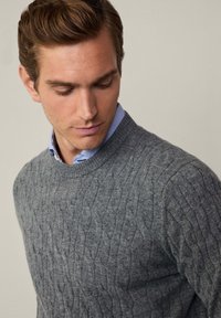 Grauer Kabelstrickpullover aus weichem Stoff, mit Rundhalsausschnitt, strukturiertem Muster und über einem hellblauen Hemd mit Kragen getragen.