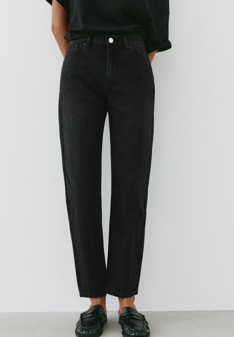 Personne portant un jean noir taille haute à jambe droite associé à des mocassins noirs et une chemise noire rentrée, debout contre un mur uni.