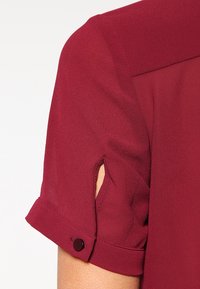 Blouse bourgogne avec manches retroussées, ornée de boutons et d'une petite fente. Fabriquée en tissu léger avec une texture douce.