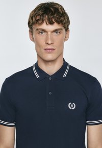 Tamsiai mėlyna polo palaidinė su apykakle, trimis mygtukais ir baltais dryžiais ant rankovių. Mažas išsiuvinėtas logotipas krūtinėje.