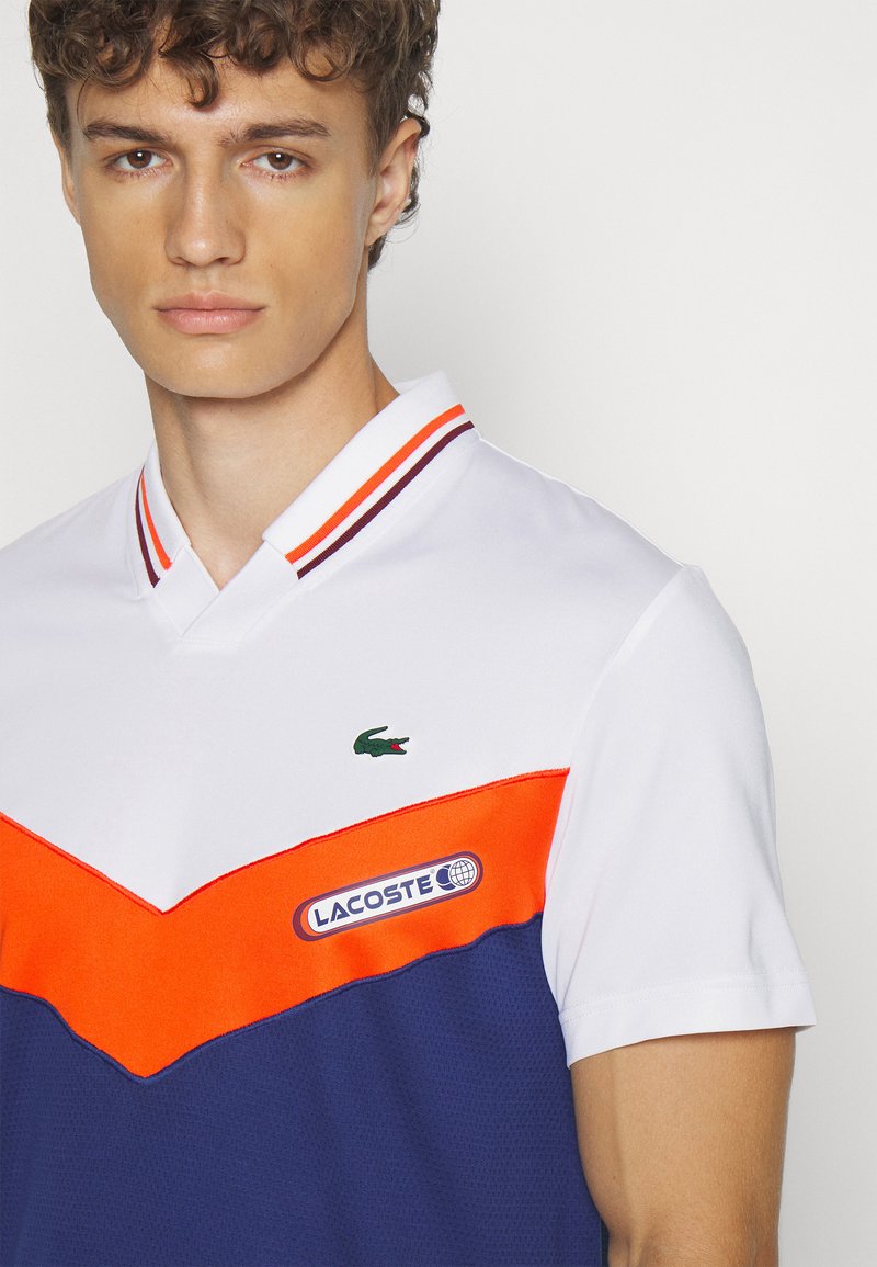 Polo à manches courtes avec un corps blanc, un motif en chevron orange et bleu marine, le logo Lacoste, et un col rayé. Tissu respirant.