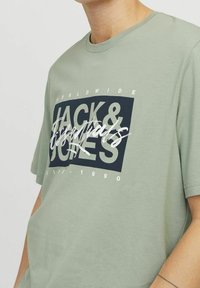 Camiseta de algodón verde oliva con un gráfico audaz en azul marino y texto blanco. Diseño sencillo de cuello redondo, mangas cortas y ajuste relajado.