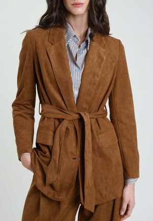 Donna che indossa un blazer color cuoio in suede con cintura sopra una camicia a righe abbottonata, con una mano in tasca, su uno sfondo chiaro e semplice.