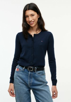 Frau lächelt, trägt eine marineblaue, geknöpfte Strickjacke mit kleinem Logo, kombiniert mit hellblauen Jeans und einem schwarzen Gürtel vor neutralem Hintergrund.