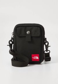 Mala tiracolo de tecido preto com fecho de correr, apresentando uma aba frontal, um botão de pressão prateado e um emblema vermelho com logótipo. Inclui uma alça ajustável.