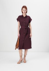 Burgundy skjortklänning med korta ärmar, knapplist fram, knytmidja och två framfickor. Matchad med beigefärgade sandaler med remmar och en brun väska.