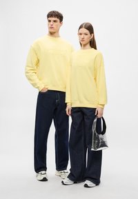 Κίτρινες βαμβακερές φούτερ με ανάγλυφο λογότυπο, σε συνδυασμό με σκούρο μπλε τζιν παντελόνια με φαρδιά γραμμή. Η γυναίκα κρατά μια διαφανή τσάντα με μαύρη λαβή.