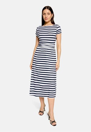 Kvinde iført en midi-kjole med korte ærmer, navy og hvide vandrette striber, med sorte strop-sandaler med høje hæle, stående mod en ensfarvet baggrund.