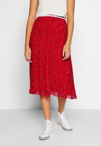 Roter plissierter Rock mit Paisleymuster, kombiniert mit einem kurzen Oberteil. Getragen mit weißen High-Top-Sneakern.