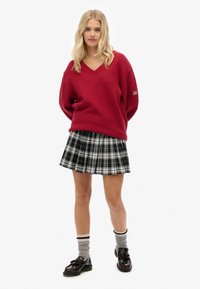 Una donna indossa un maglione rosso a V, una gonna a pieghe a scacchi bianca e nera, calze grigie con righe e mocassini neri con nappine.