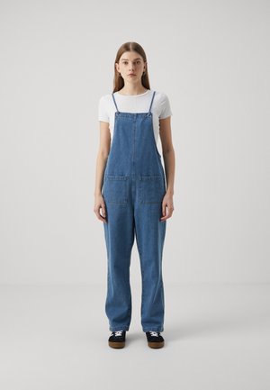 Jeansoverall i medelblått med justerbara axelremmar, två framfickor och vida ben. Matchas med en tajt vit skjorta och svarta sneakers.