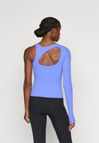 Ελαφρύ μωβ αθλητικό τοπ με ένα μακρύ μανίκι και ανοιχτή πλάτη, με λογότυπο Nike κοντά στο τελείωμα. Συνδυάζεται με μαύρο κολάν.