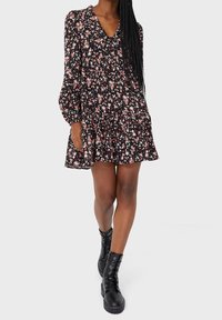 Zwarte bloemenjurk met lange mouwen, V-hals en gerimpelde zoom. Bevat kleine roze en witte bloemen op een donkere achtergrond. Draagt met zwarte combatboots.