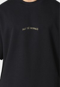 Black cotton t-shirt featuring short sleeves and a round neckline, with "PAS DÉ MONACO" embroidered in gold above the chest.