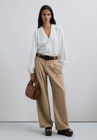 Blusa blanca de manga larga con cuello fruncido, pantalones beige de pierna ancha, cinturón marrón, bolso marrón y zapatos de plataforma oscuros.