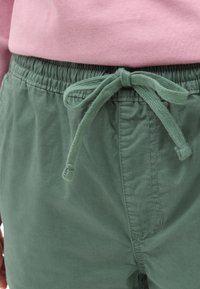 Pantalones cortos verdes con cintura elástica y cordón, fabricados en suave algodón. Presentan una textura lisa y detalles de costura visibles.