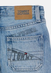 Jeans di jeans azzurro chiaro con una toppa in pelle marrone impressa "TOMMY HILFIGER." Presentano una tasca ricamata con un piccolo logo in rosso, bianco e blu.