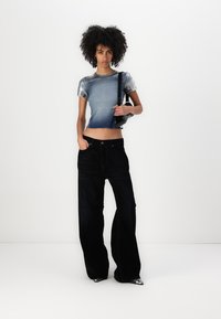 Metallisk sølv kortermet topp med en gradient blå design, kombinert med vide svarte jeans. Modellen holder en liten svart veske.