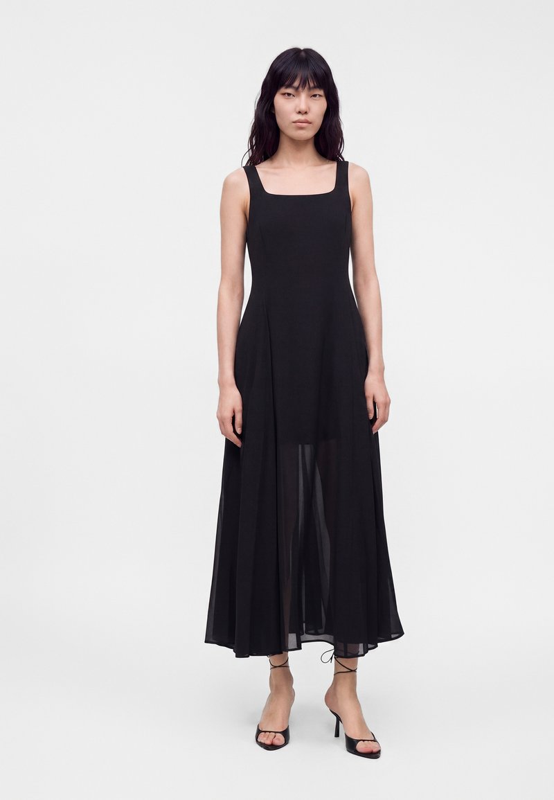 Femme aux longs cheveux foncés portant une robe midi noire sans manches avec un décolleté carré et des talons noirs à brides, debout sur un fond uni.