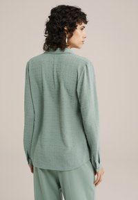 Helle grüne Bluse aus texturiertem Stoff mit kleinen, erhabenen Mustern, langen Ärmeln und einem schlichten Kragen.