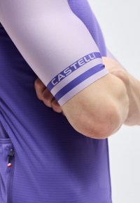 Κοντινό πλάνο λυγισμένου βραχίονα με ανοιχτό μωβ μανίκι ποδηλασίας που φέρει την επιγραφή "CASTELLI" και κοντομάνικο μπλουζάκι σκούρου μοβ χρώματος.