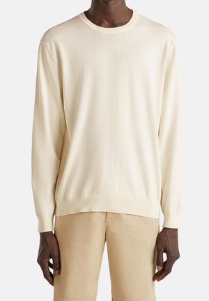 Homme portant un pull crème uni à manches longues et un pantalon beige, debout sur un fond blanc.