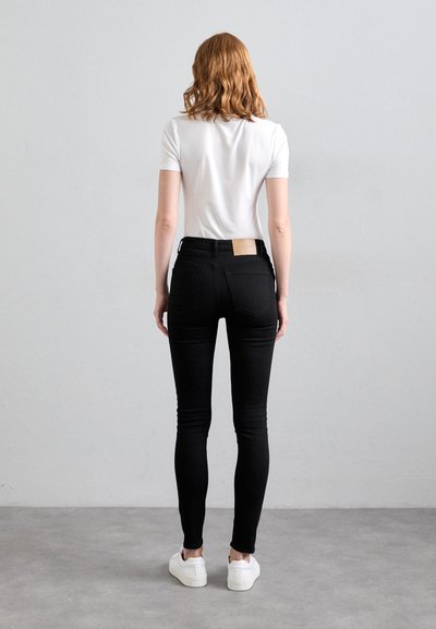 Schwarze, enge Jeans mit hoher Taille, ausgestattet mit Gesäßtaschen und einem hellbraunen Lederlabel. Kombiniert mit einem kurzärmeligen weißen T-Shirt und weißen Sneakers.