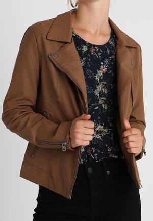 Kvinde iført en brun lynlåstrøje over en mørk blomstret bluse, som holder jakkeenderne med begge hænder mod en ensfarvet baggrund.