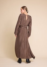 Lang, brun satindress med lange ermer, detalj med nøkkelhull i ryggen og belte i midjen. Glatt tekstur og flytende silhuett. Svarte ankelstøvler.