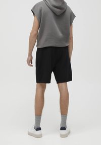 Sweat à capuche sans manches gris avec une capuche, associé à un short noir. Le short possède une taille élastique, accompagné de baskets blanches et de chaussettes grises.