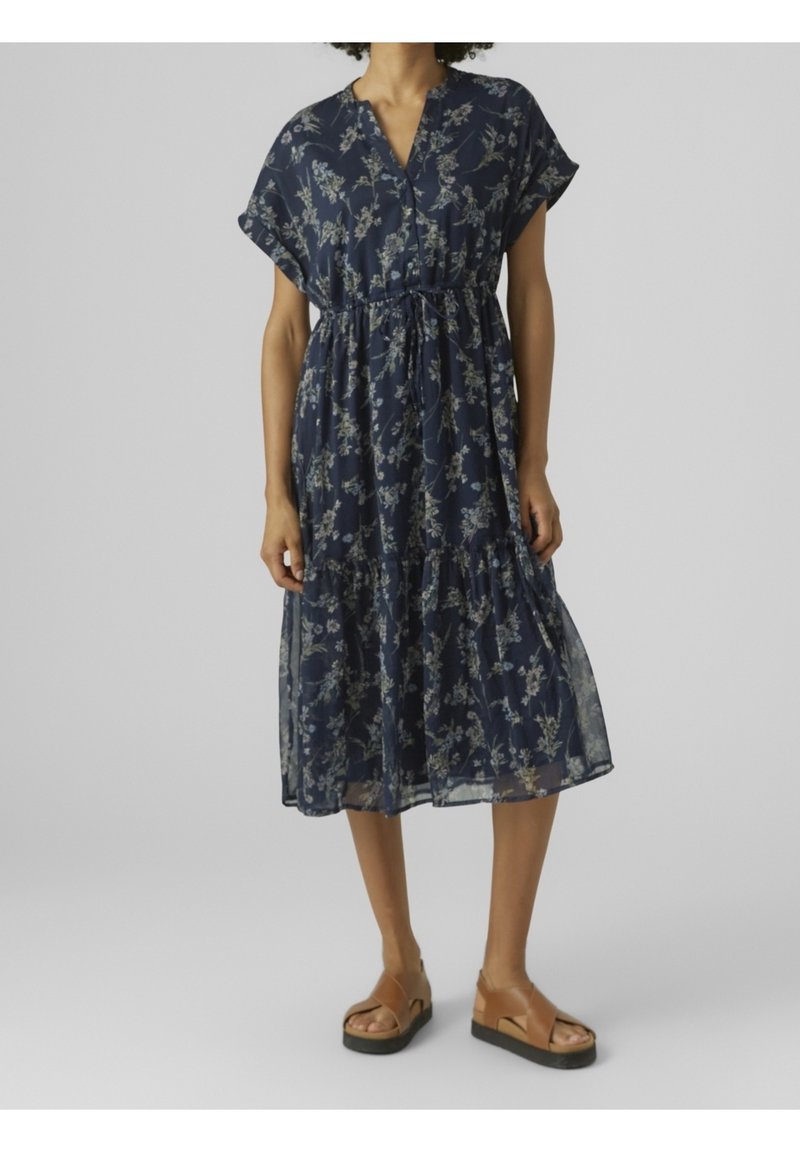 Femme portant une robe midi fleurie bleu marine à manches courtes et des sandales compensées marron, debout devant un fond clair uni.