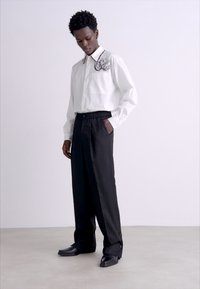White long-sleeve shirt with embroidered design at the collar, paired with black wide-leg trousers and black shoes. Smooth fabric texture.