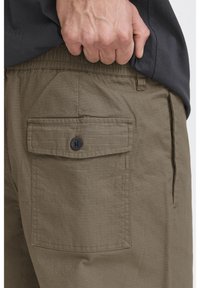 Khaki cargo shorts with a rectangular back pocket featuring a black button. The fabric has a textured, grid pattern. Elasticated waistband.