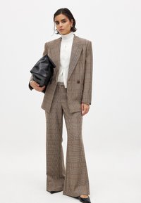 Geverifieerde pakset in bruintinten, met een dubbelgeknoopte blazer en wijde broek. G gedragen met een witte turtleneck en een zwarte tas.