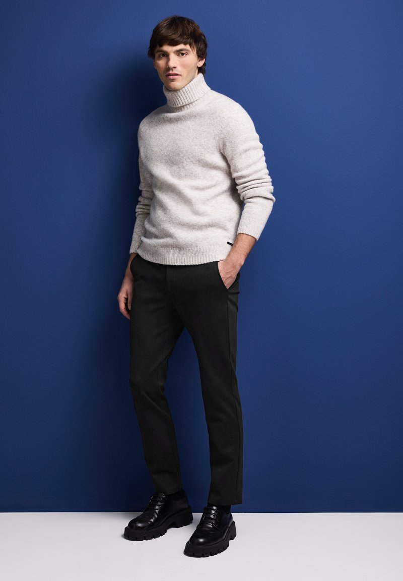 Ljust grått polotröja, svarta slim-fit byxor, svarta skor med kraftig sula. Modellen står mot en blå vägg, med händerna i byxfickorna.